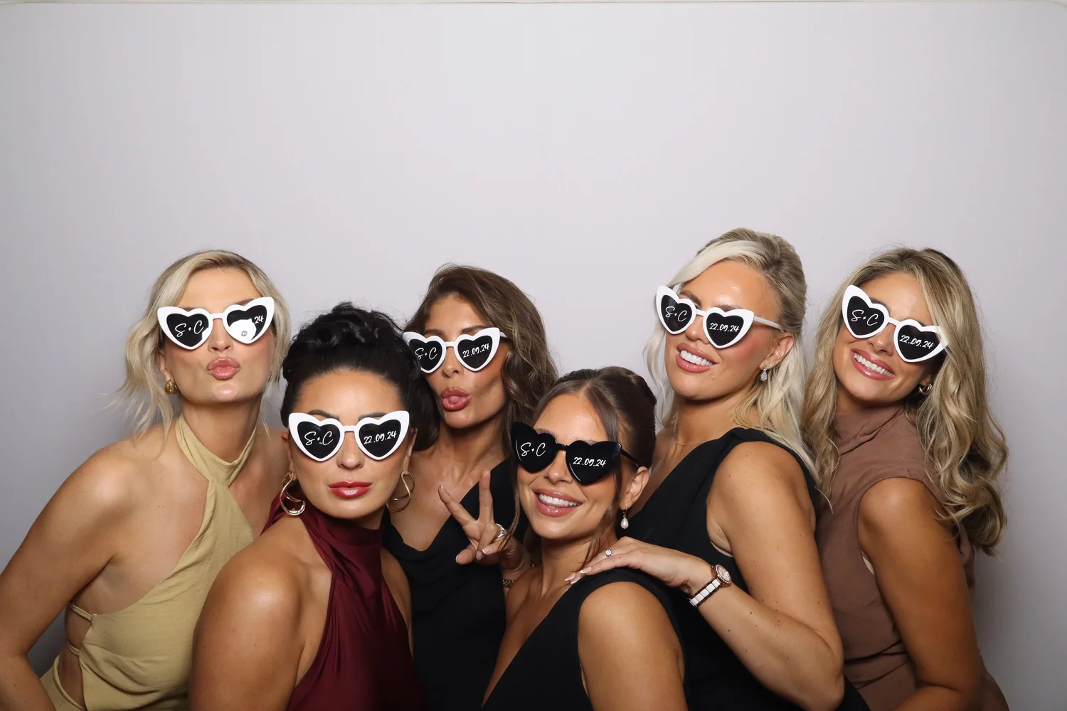 Group of seven women celebrating, wearing heart-shaped sunglasses with 'S+C' and a date written on them, posing playfully against a plain background.