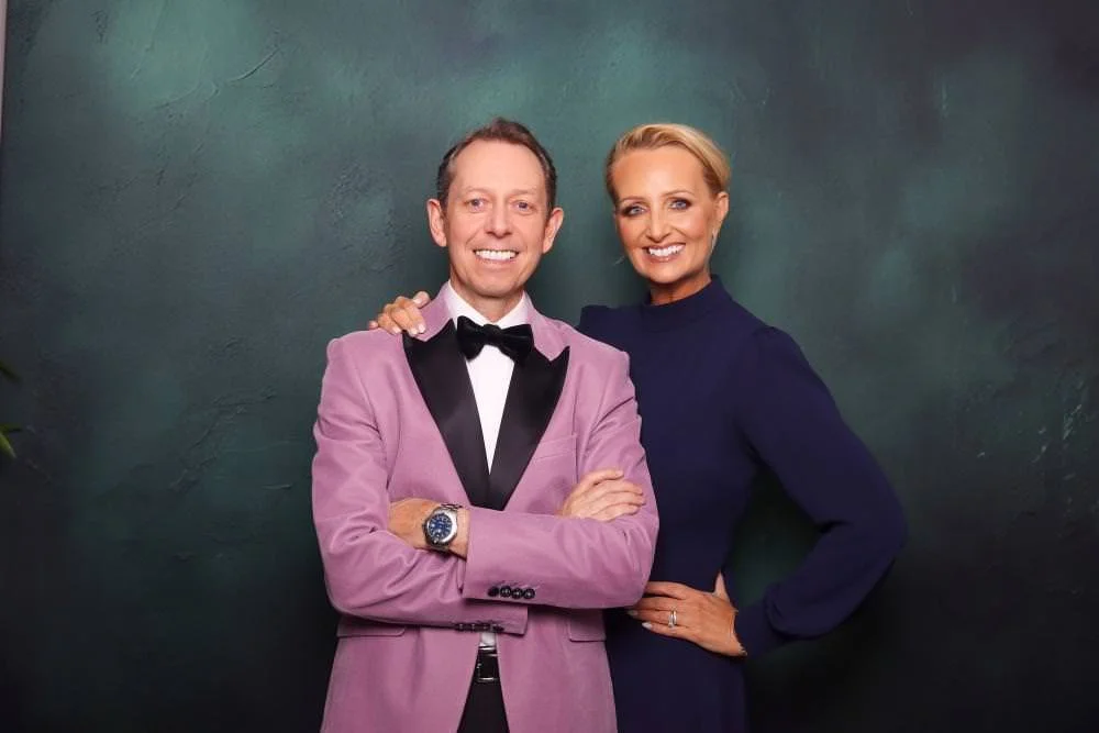 A man and woman posing together in front of a green backdrop. The man is wearing a pink tuxedo with a black bow tie and a white shirt, and the woman is wearing a dark blue dress. The man has his arms crossed and is smiling, while the woman has her ha