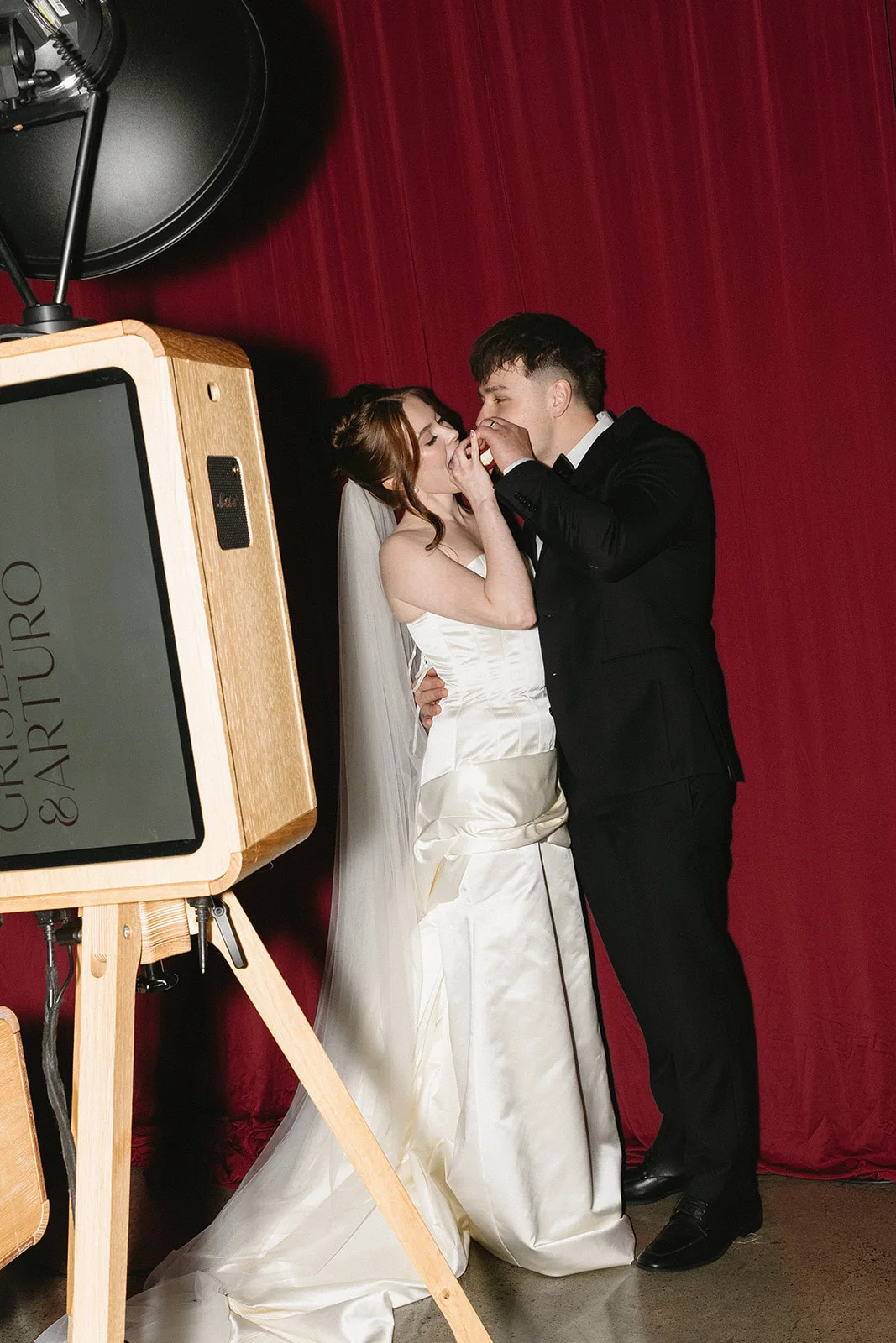 Bride and groom beside the wooden ICON photo booth in front of a red curtain