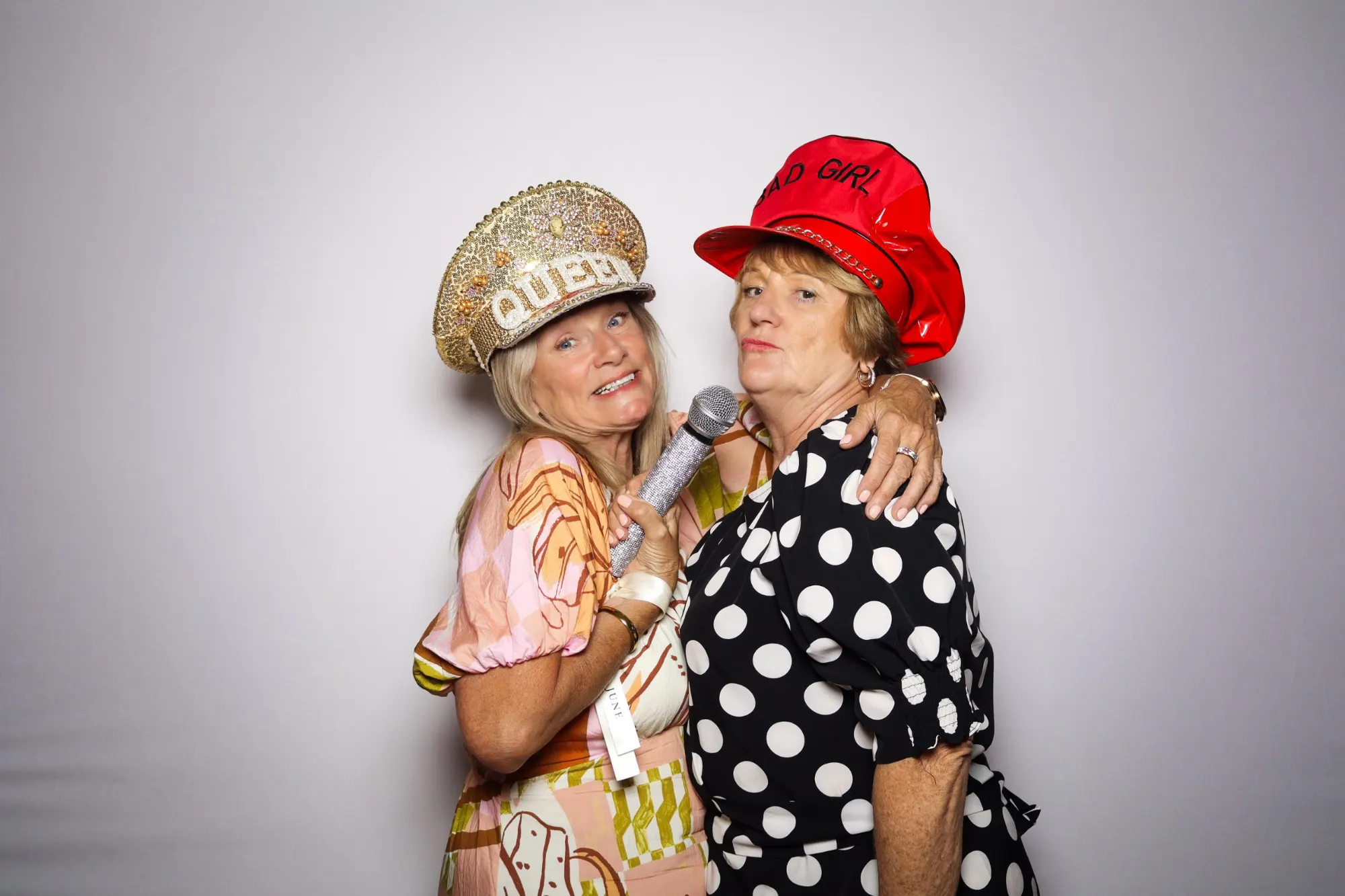 Two wedding guests in a sparkly Queen hat and a red cap posing in the photo booth