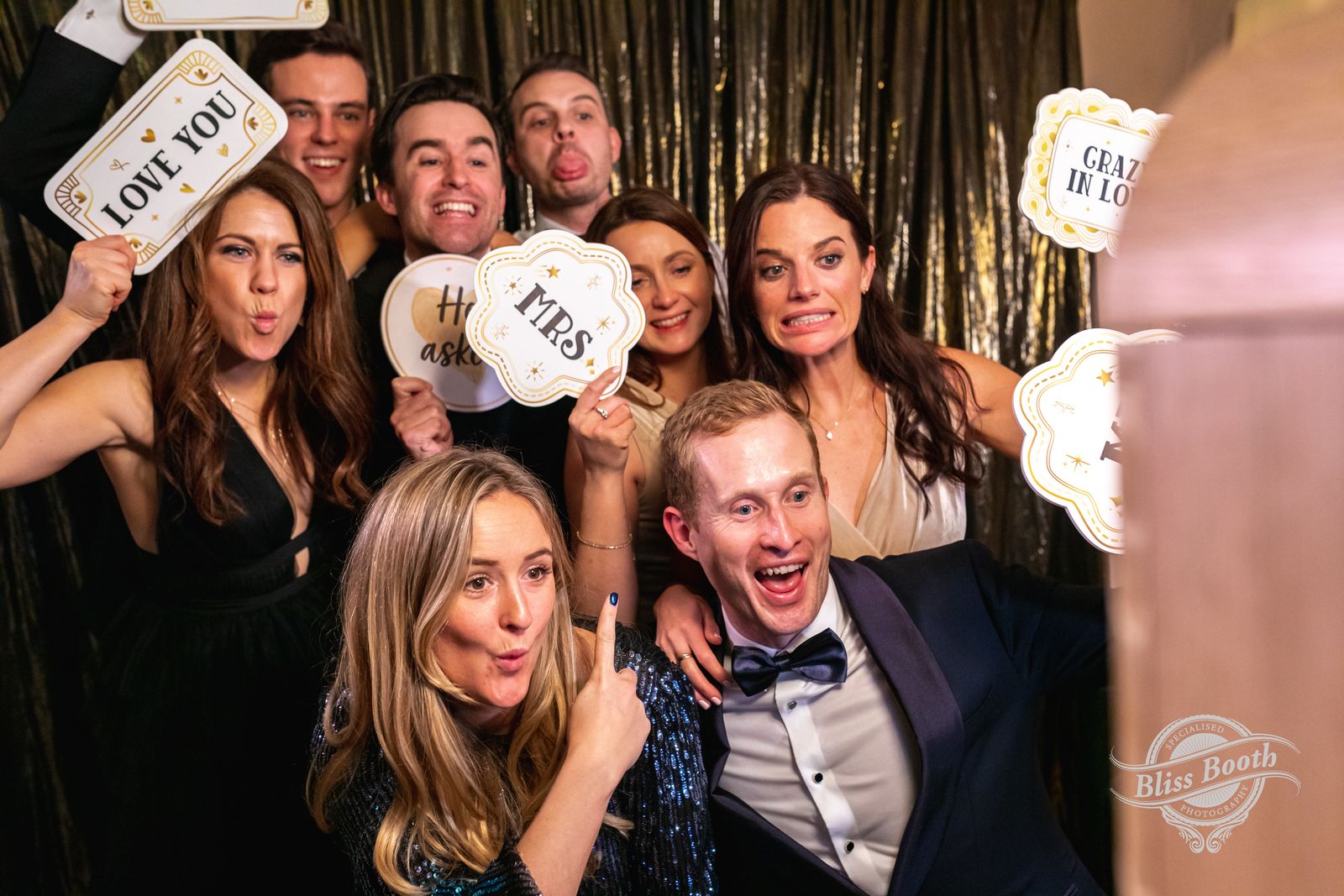 Wedding guests laughing with signs in front of a gold tinsel photo booth backdrop