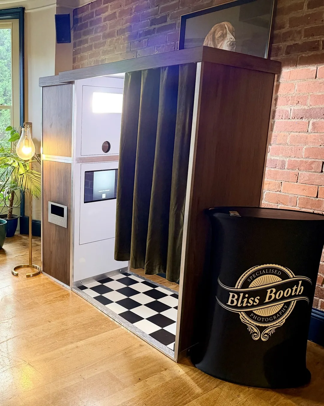 Wooden Photomatic photo booth with checkerboard floor set up beside a brick wall at a wedding venue