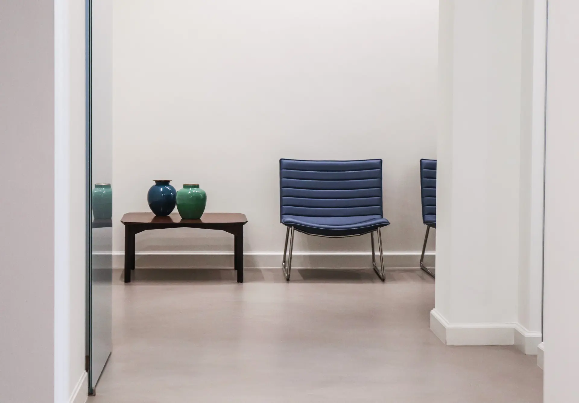 Minimalist interior with a blue cushioned chair with metal legs and a wooden side table holding two decorative vases, one dark blue and one green, against a plain light-colored wall.