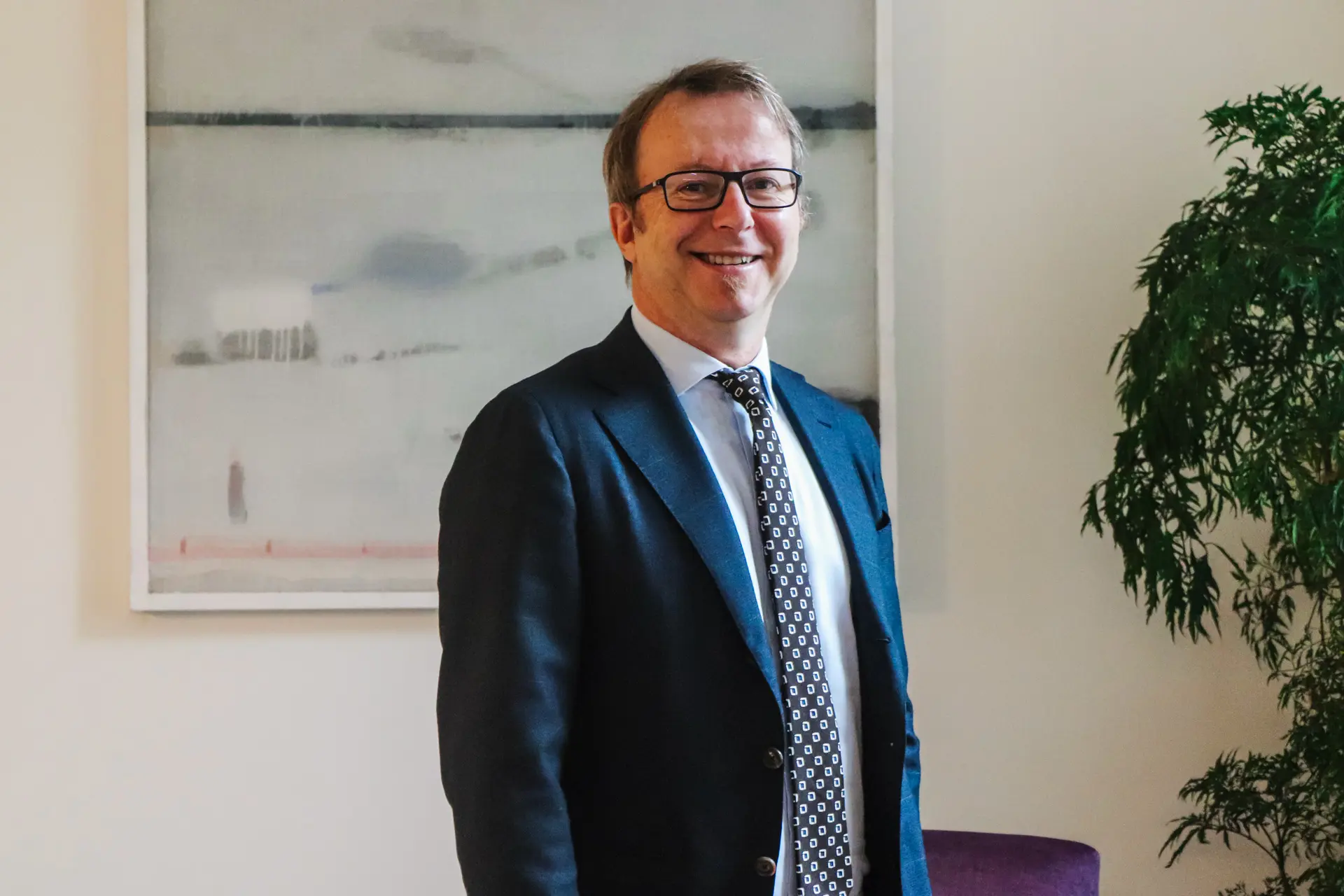Smiling man with glasses wearing a dark suit, white shirt, and patterned tie standing indoors with abstract art and a green plant in the background.