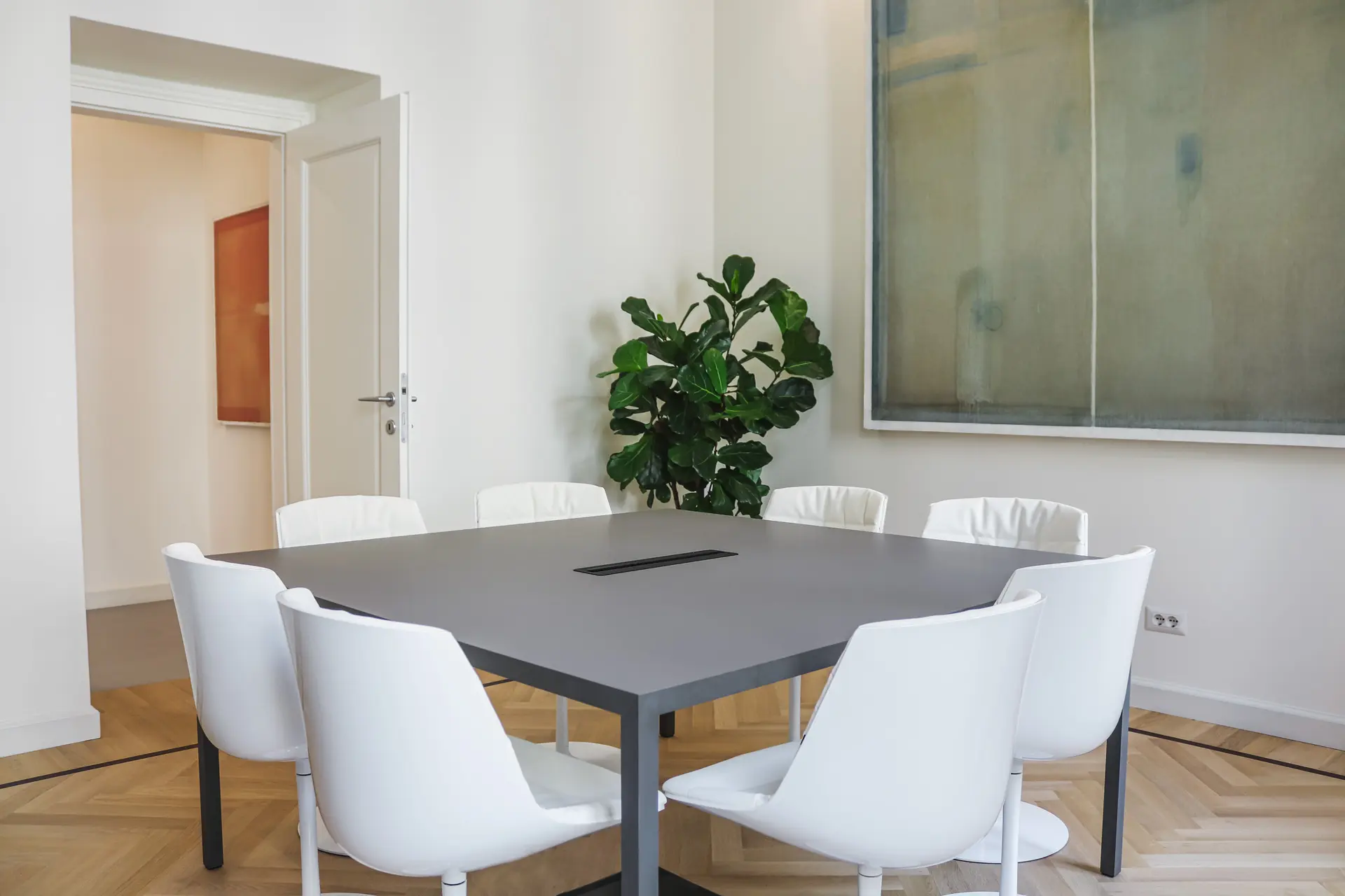 Modern meeting room with a dark square table surrounded by eight white chairs, a green leafy plant in the corner, light-colored wood herringbone floor, and a large abstract painting on the wall.