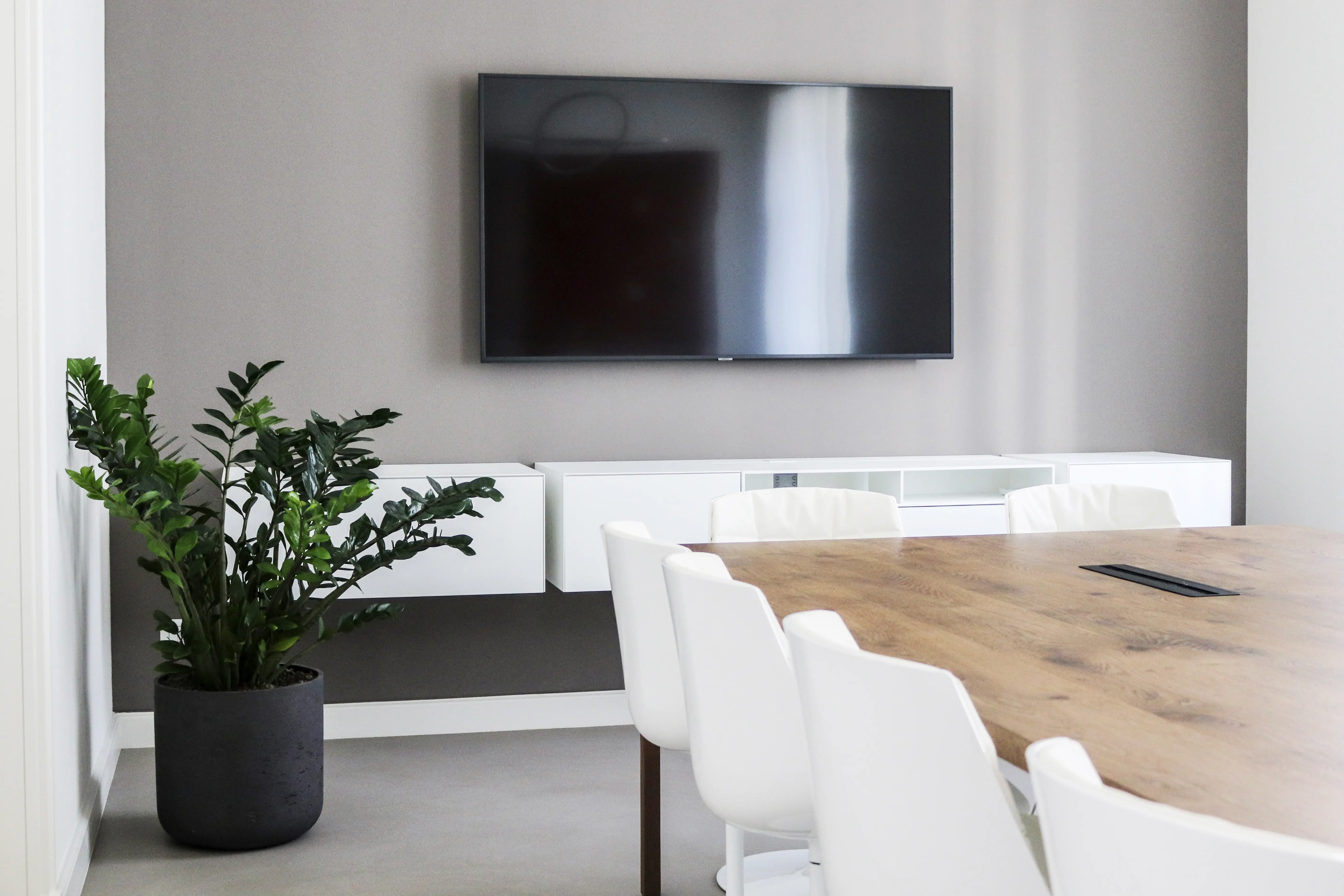Modern office meeting room with a large wooden table surrounded by white chairs, a flat-screen TV mounted on a gray wall, white floating cabinets, and a green potted plant in a dark pot on the floor.