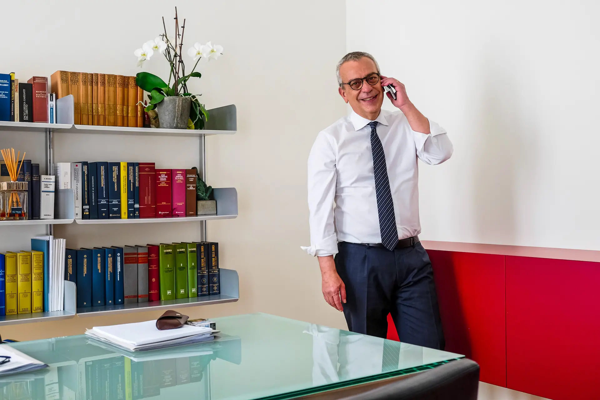 Smiling middle-aged man in glasses, white shirt, and tie talking on a smartphone, leaning against a red and white wall in a modern office with a glass table and a bookshelf filled with colorful books and a potted orchid.