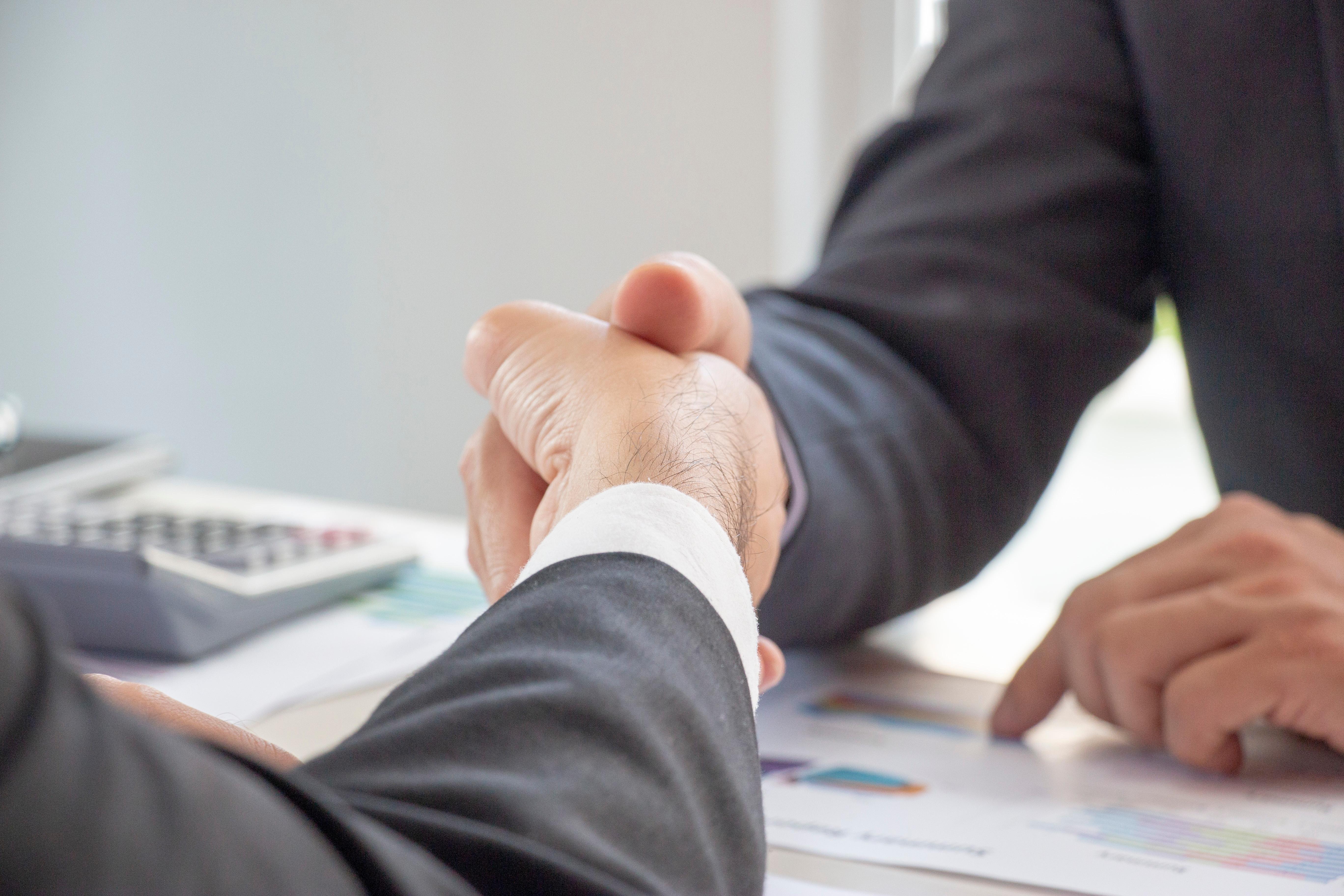 Two people in business suits shaking hands over a desk with documents and a calculator, symbolizing a business agreement or partnership.