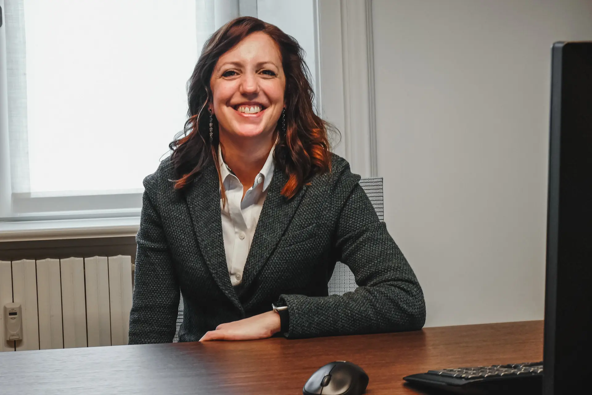 Smiling woman with wavy brown hair wearing a dark textured blazer and white shirt, sitting at a desk with a computer keyboard and mouse.