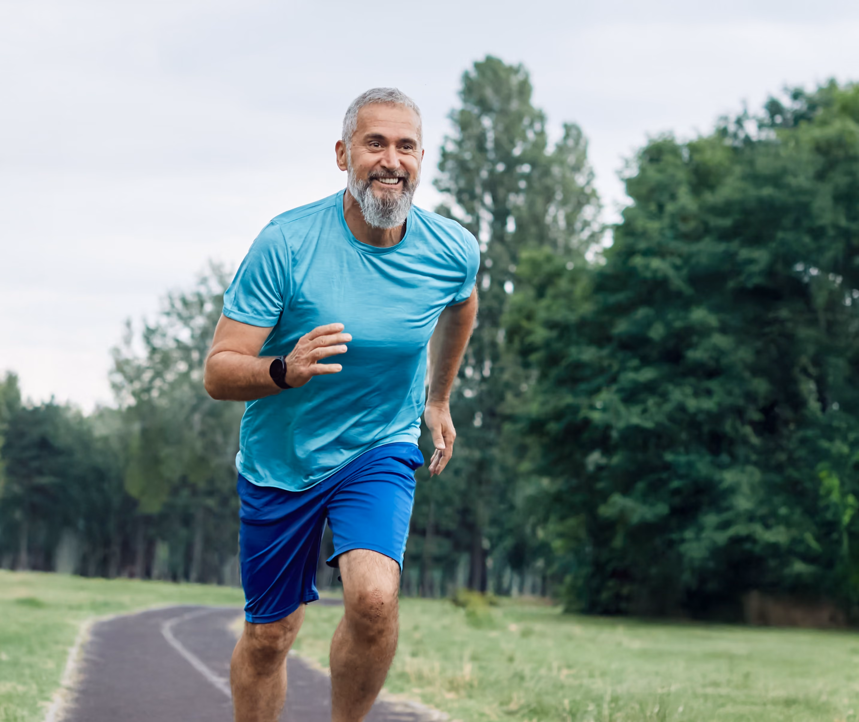 Middle-aged man with gray hair and beard running on a paved path in a park, wearing a turquoise shirt and blue shorts, smiling with trees and grass in the background.