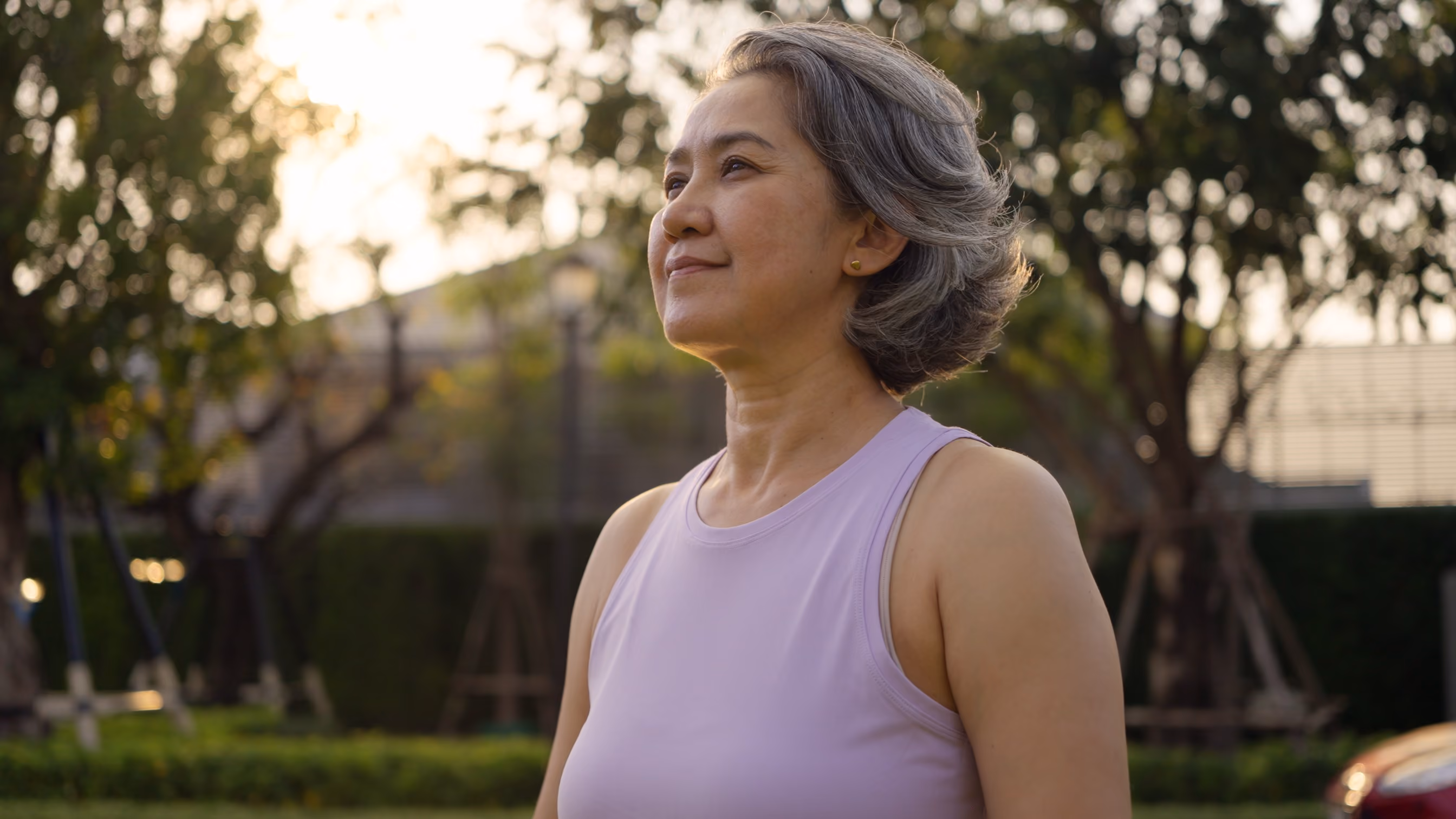 Middle-aged woman with short gray hair wearing a sleeveless lavender top standing outdoors in a park during sunset, looking forward with a calm and content expression.