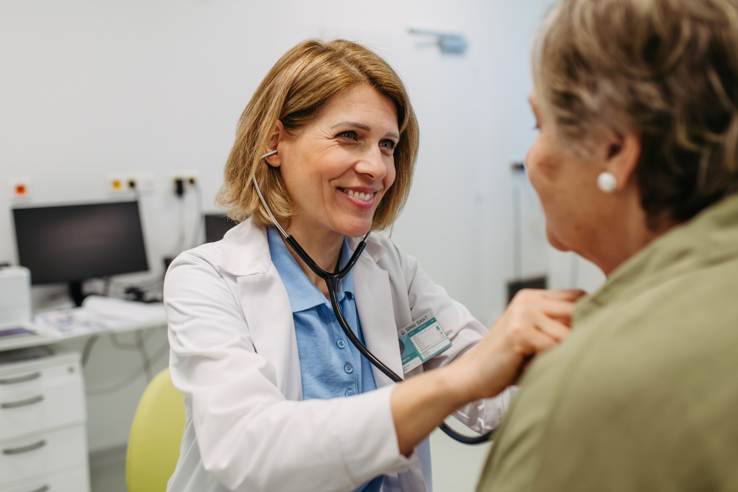Female doctor wearing a white coat and blue shirt using a stethoscope to examine a patient in a medical office.