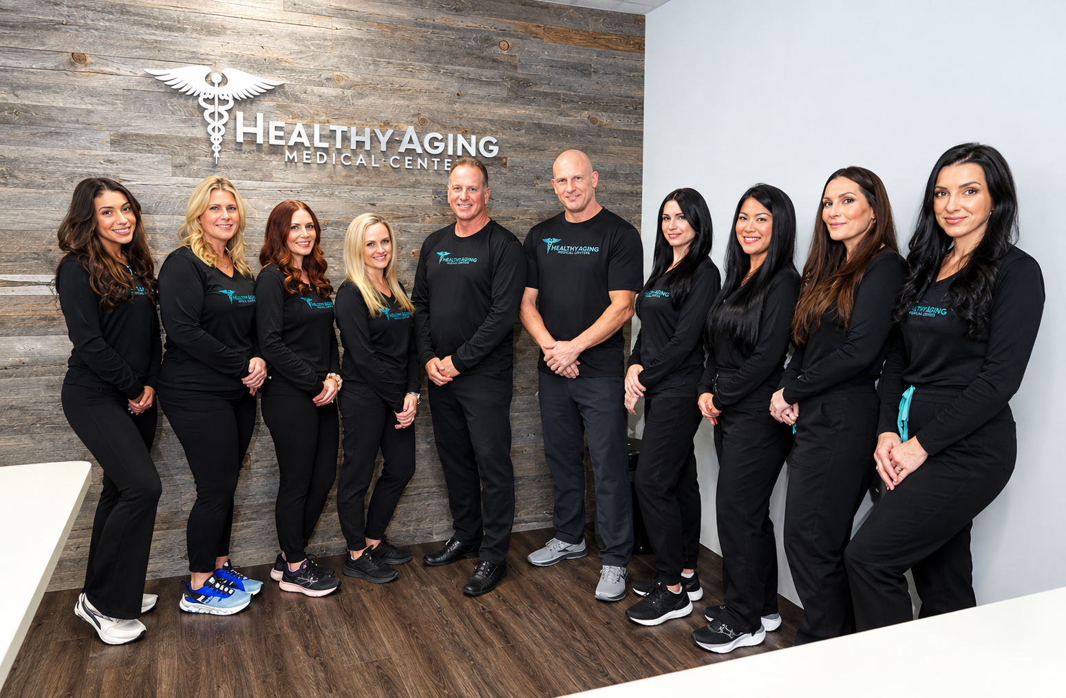 A group of ten medical professionals, eight women and two men, standing side by side in a reception area with a wood-paneled wall behind them displaying the logo and name Healthy Aging Medical Center. All are wearing matching black uniforms with the medical center's logo.