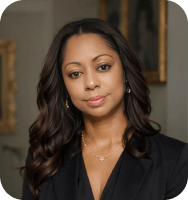 Portrait of a woman with long wavy dark hair wearing a black blazer and delicate gold jewelry, looking confidently at the camera with a neutral expression, against a softly blurred indoor background.