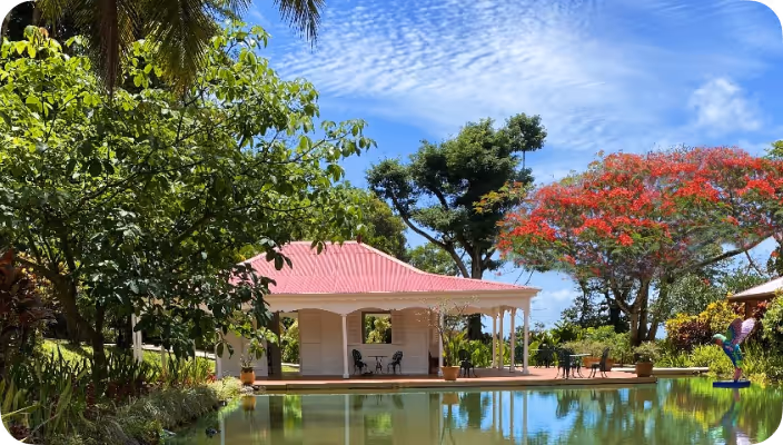 Petit pavillon blanc avec toit rose entouré d’arbres verts et d’un arbre à fleurs rouges au bord d’un étang calme reflétant le paysage.