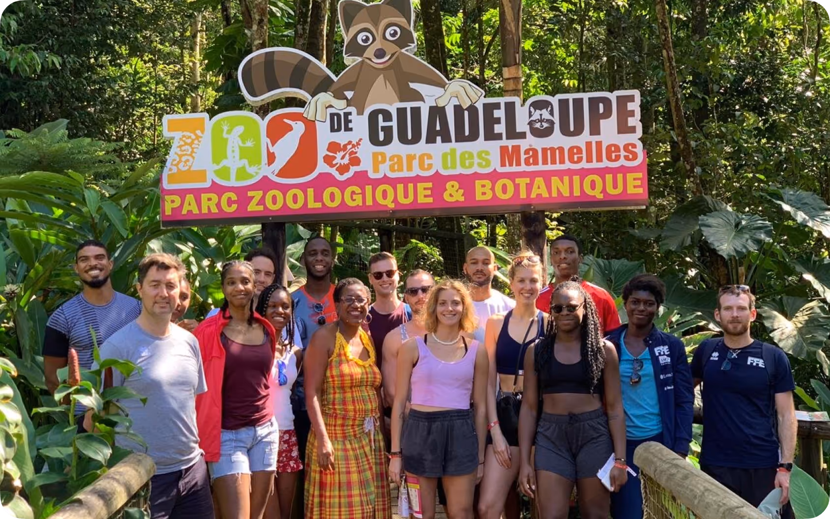Un groupe de personnes posant devant le panneau coloré du Zoo de Guadeloupe au Parc des Mamelles entouré de végétation luxuriante.