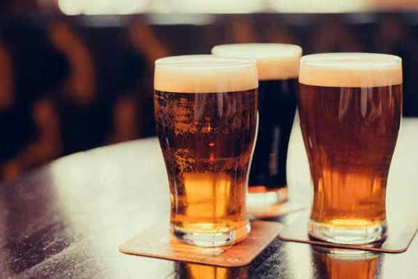 Three pint glasses of beer with foam heads on a wooden table, with warm lighting and a blurred background.