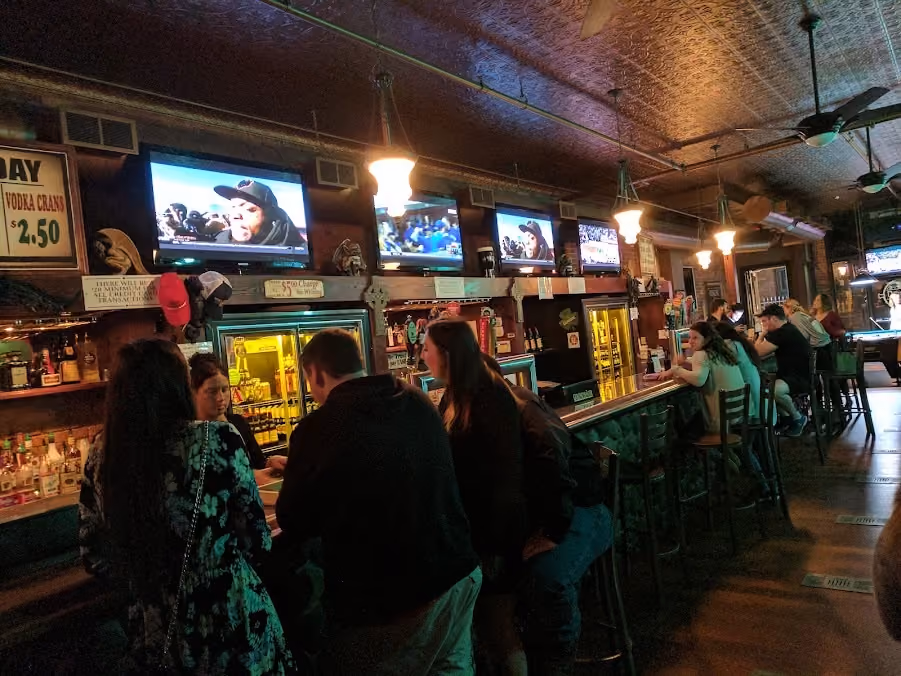 Dimly lit bar interior with people sitting and conversing at the long wooden bar counter with green padded seating. Multiple TV screens above the bar show different programs. Pendant lights hang from the decorative ceiling. Shelves behind the bar display liquor bottles and hats, with signs advertising drink prices.