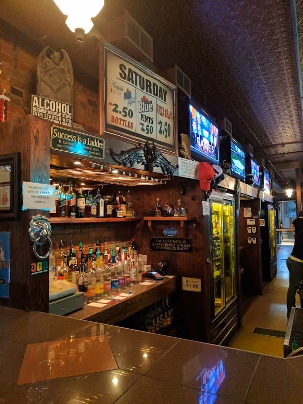 Interior of a cozy bar with a wooden counter and shelves stocked with bottles of liquor and mixers, a fridge with drinks, multiple TV screens on the wall displaying sports, and various humorous and promotional signs including one advertising Saturday drink specials for Michelob Ultra, Blue, and PBR priced at $2.50, and signs emphasizing customer rights and success.