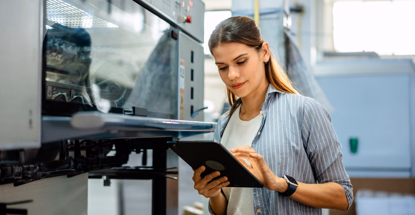 Young woman in a striped shirt using a tablet device while standing next to industrial machinery in a brightly lit workshop or factory.