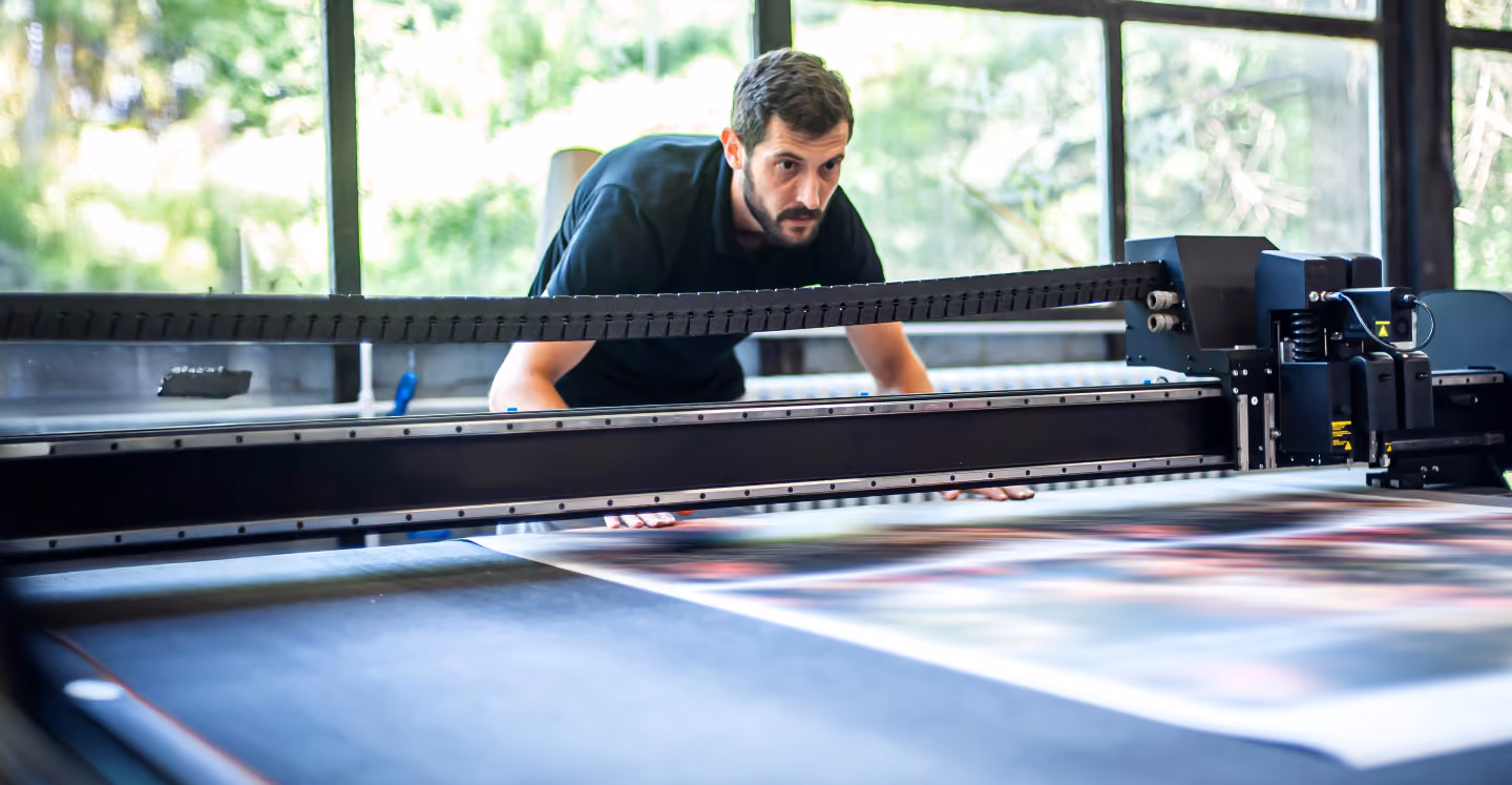 Man closely inspecting a large flatbed printer producing a wide, colorful print in a bright room with large windows.