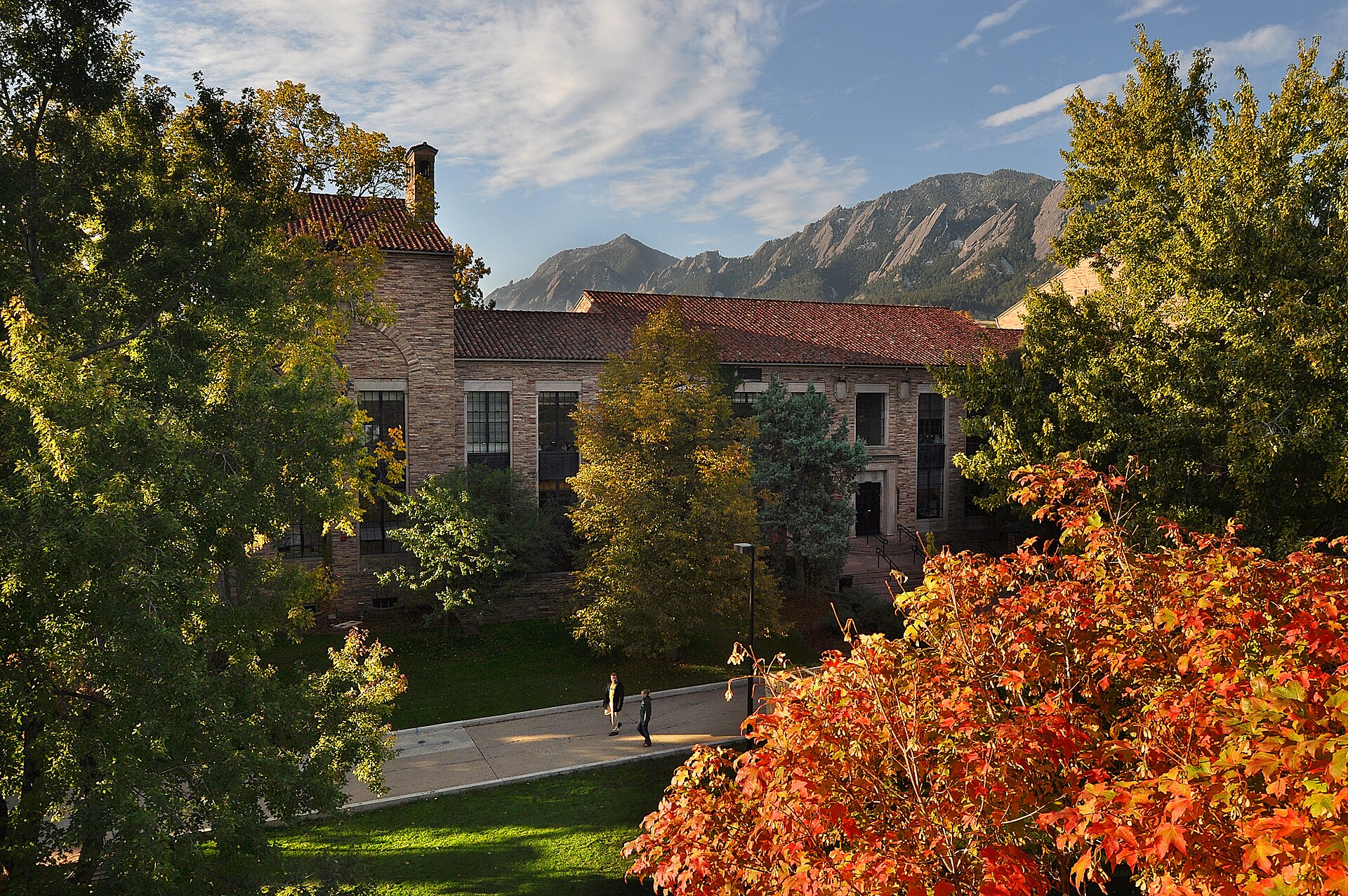 Students walking past a CU Boulder building in autumn with the Flatirons behind
