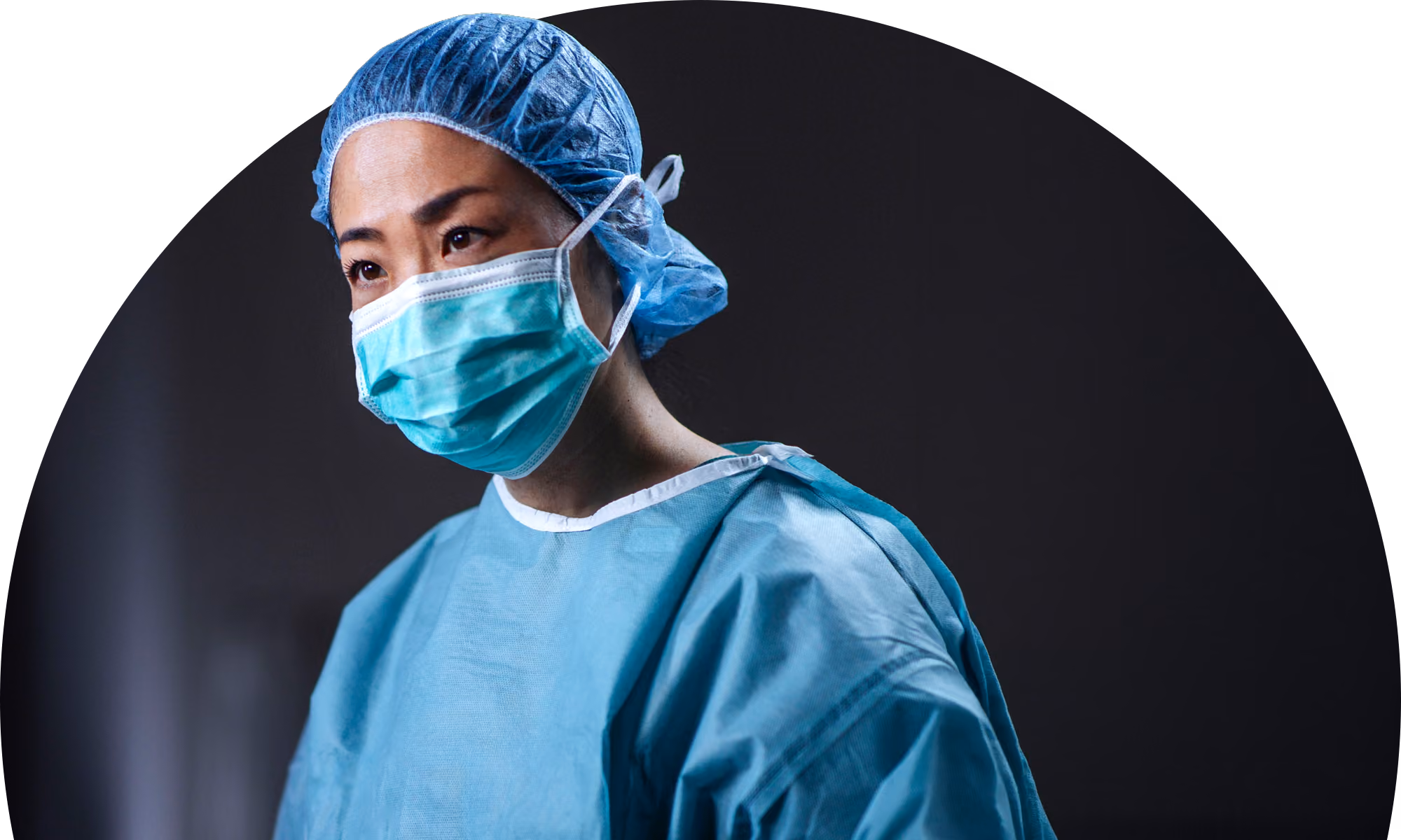 Focused healthcare professional wearing a blue surgical gown, disposable bouffant cap, and face mask against a dark background.