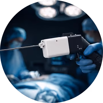 Close-up of a medical professional's gloved hand holding a white, handheld surgical device with a long thin needle in an operating room with blurred surgeons and surgical lights in the background.