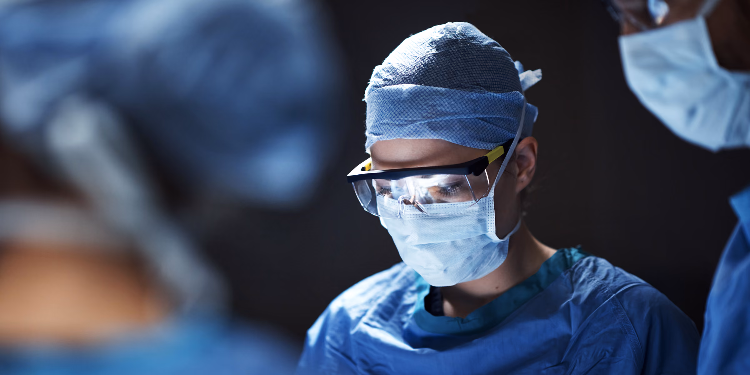 Three medical professionals wearing surgical masks, caps, and protective goggles, focused on a procedure in a dimly lit operating room.