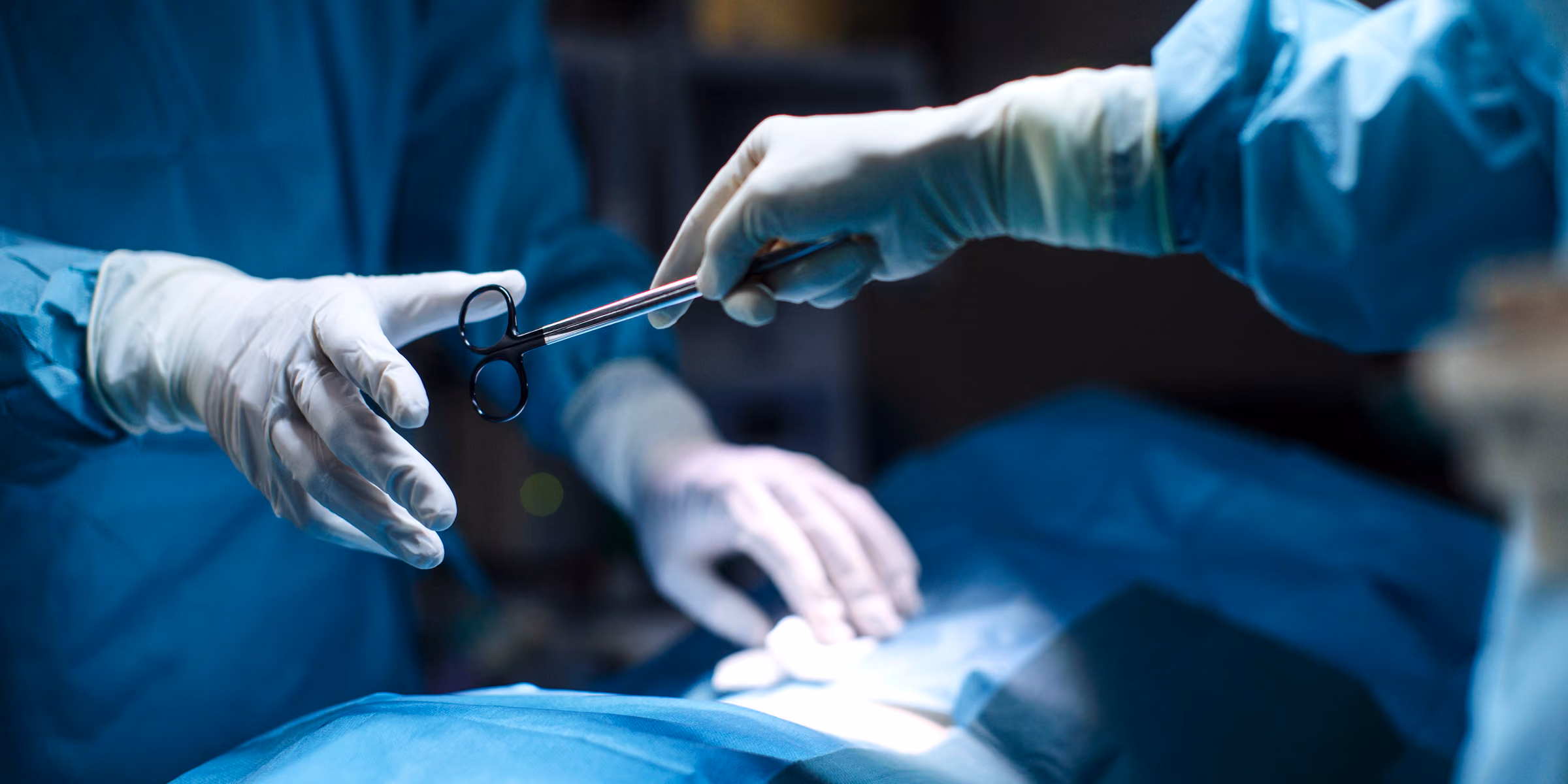 Two surgeons in blue surgical gowns and white gloves passing a surgical instrument during an operation under bright lighting.