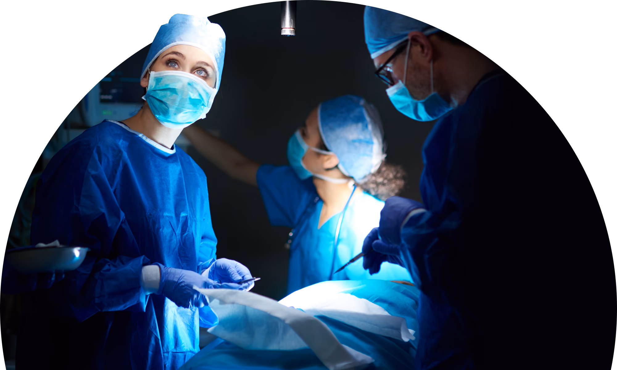 Three medical professionals in blue surgical scrubs and masks performing surgery under bright operating room lights, with one holding a surgical instrument and another preparing materials.