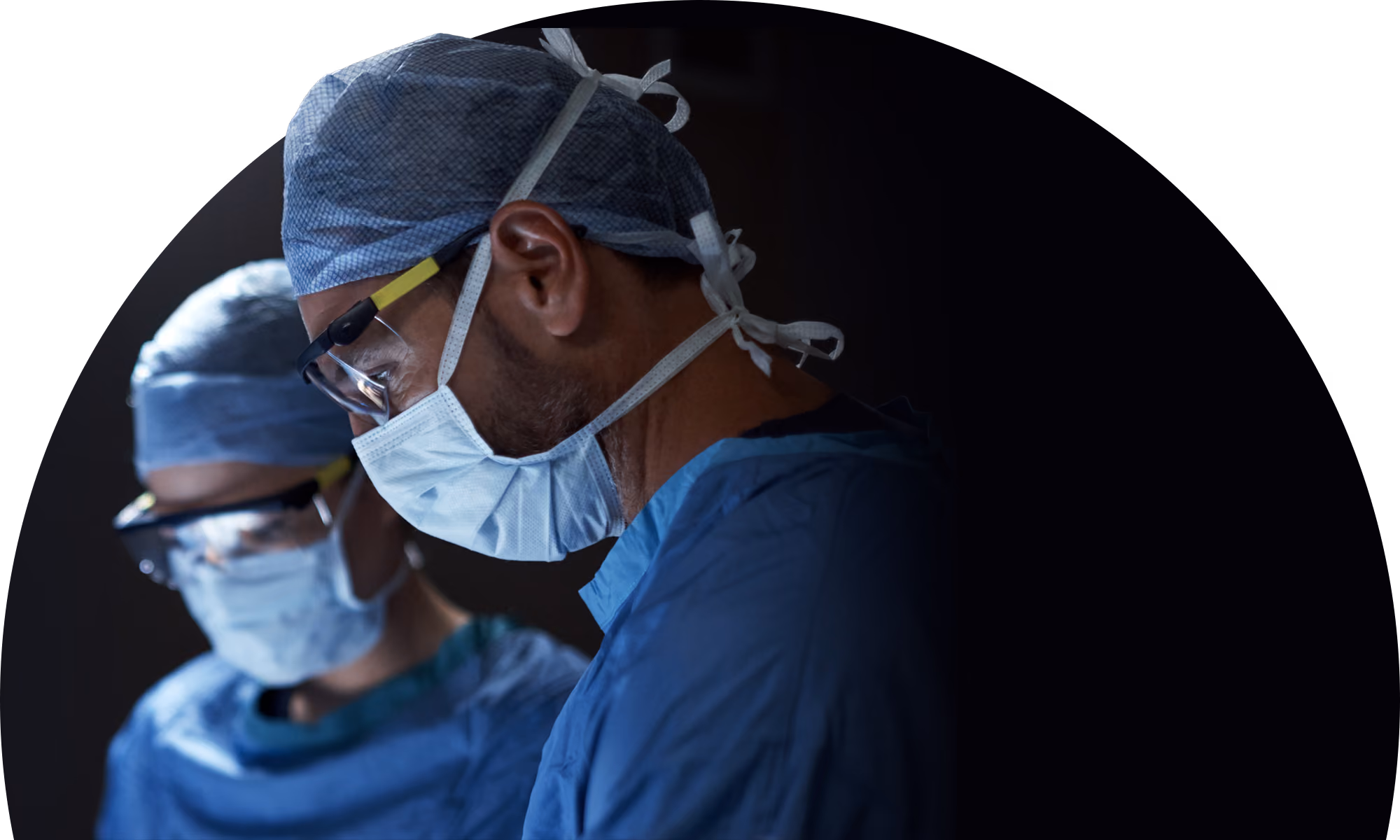 Two surgeons in blue scrubs, hair covers, face masks, and protective glasses focused on surgery in a dimly lit operating room.