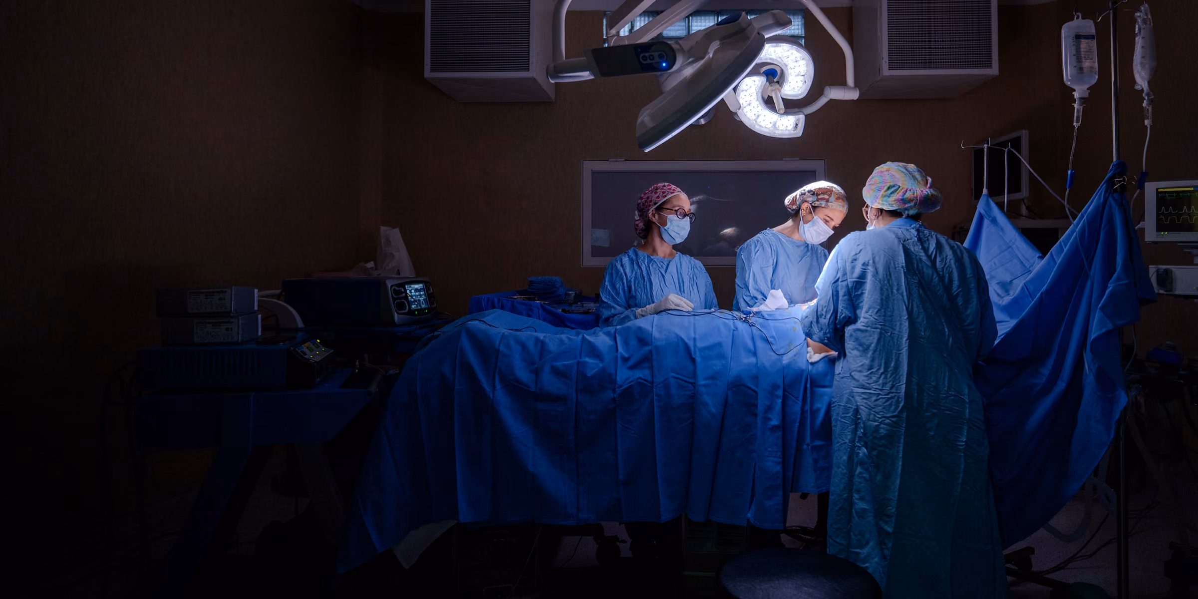 Three surgeons in blue surgical gowns and masks performing an operation under a bright surgical light in an operating room.