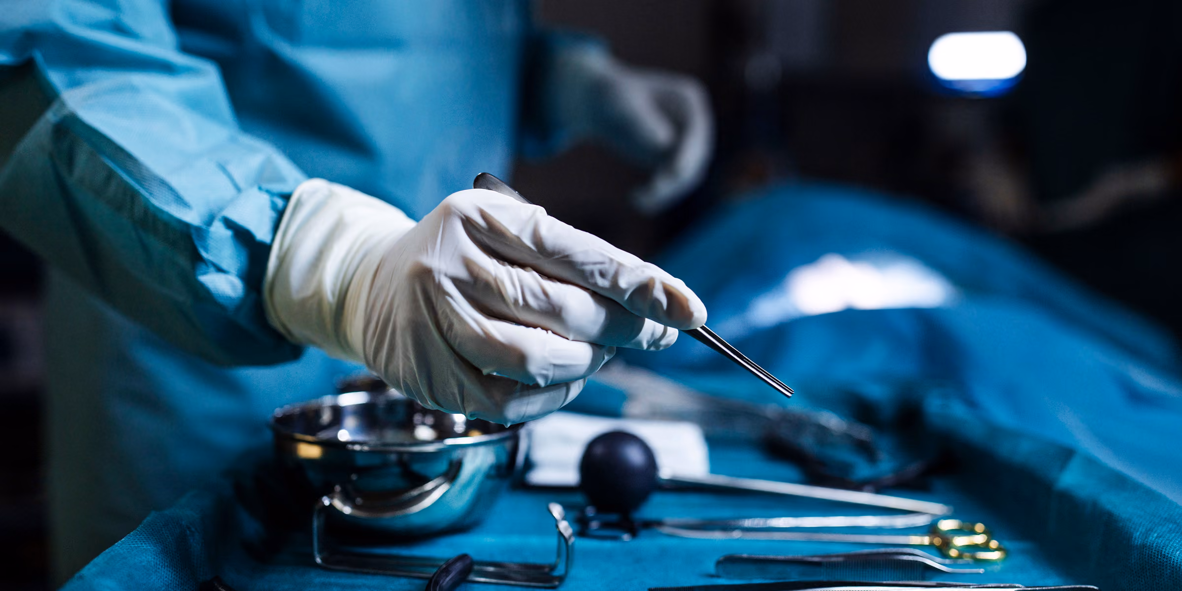 A surgeon wearing blue scrubs and white gloves holding a surgical instrument over a table with various surgical tools and a metal bowl in an operating room.