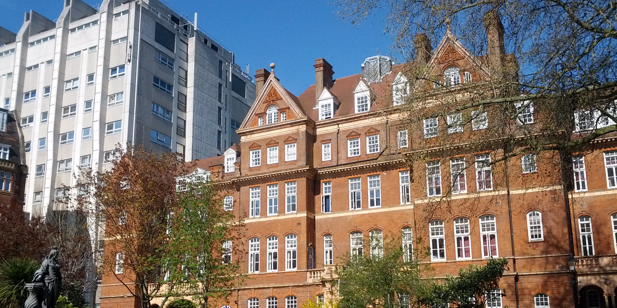A large red-brick Victorian-style building with many white-framed windows, surrounded by trees and with a modern multi-story gray building in the background under a clear sky.