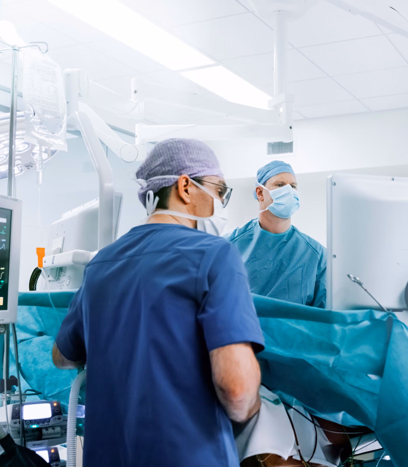 Two surgeons wearing blue surgical scrubs, caps, and face masks in an operating room, focused on a screen during surgery with monitoring equipment nearby.