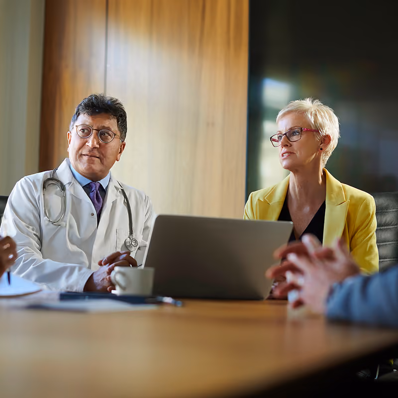 A male doctor in a white coat with a stethoscope and a woman in a yellow blazer having a conversation at a conference table with a laptop and coffee cup.