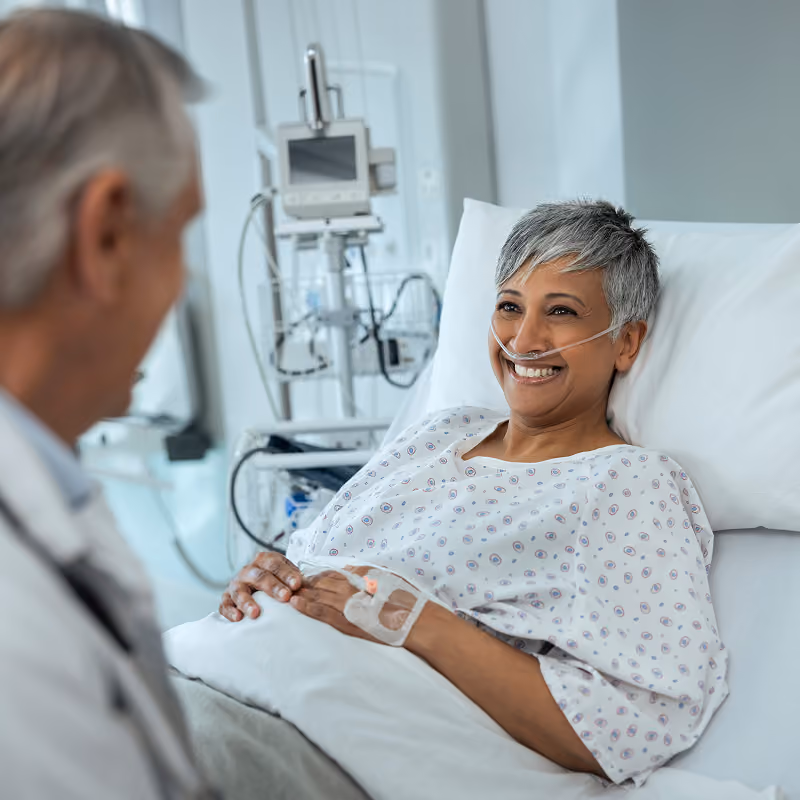 Smiling patient with short gray hair lying in a hospital bed wearing a nasal oxygen tube and hospital gown, holding hands with a doctor standing nearby.