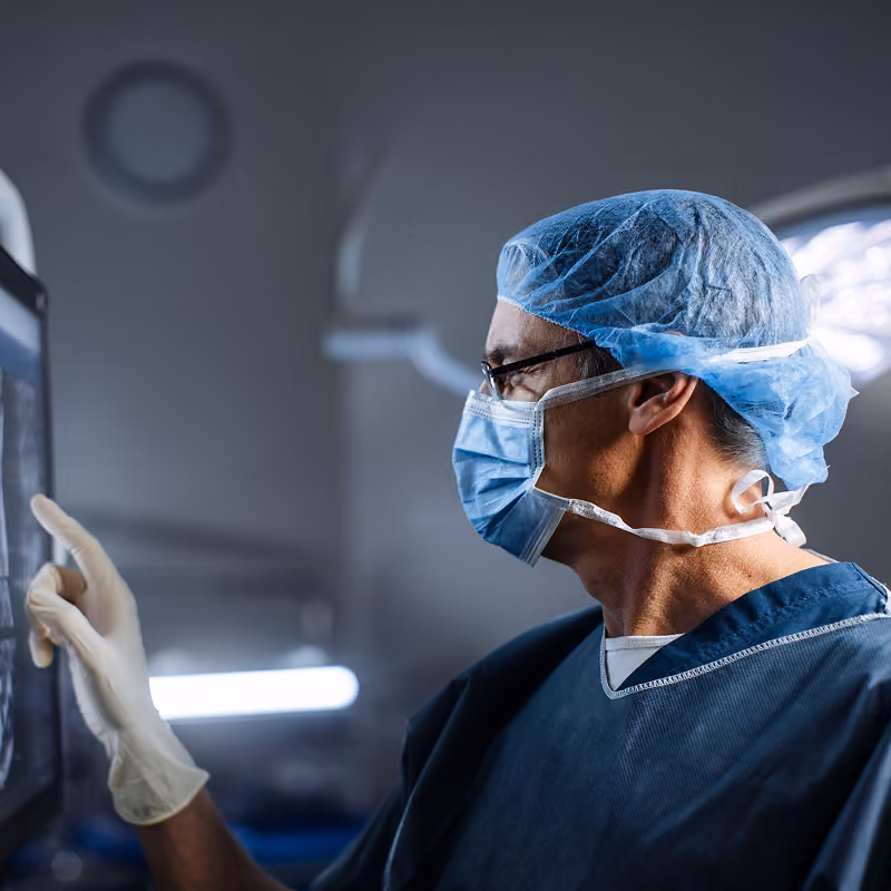 Medical professional wearing surgical cap, face mask, glasses, and gloves, reviewing an image on a touchscreen monitor in a clinical setting.
