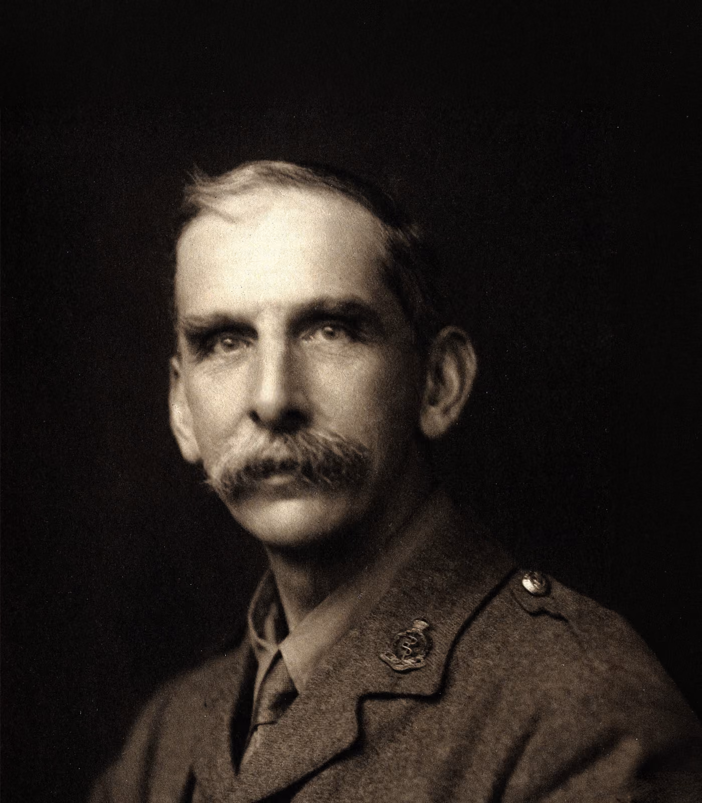 Black and white portrait of a middle-aged man with a thick mustache wearing a vintage military uniform with insignia on the shoulder and chest, looking directly at the camera against a dark background.