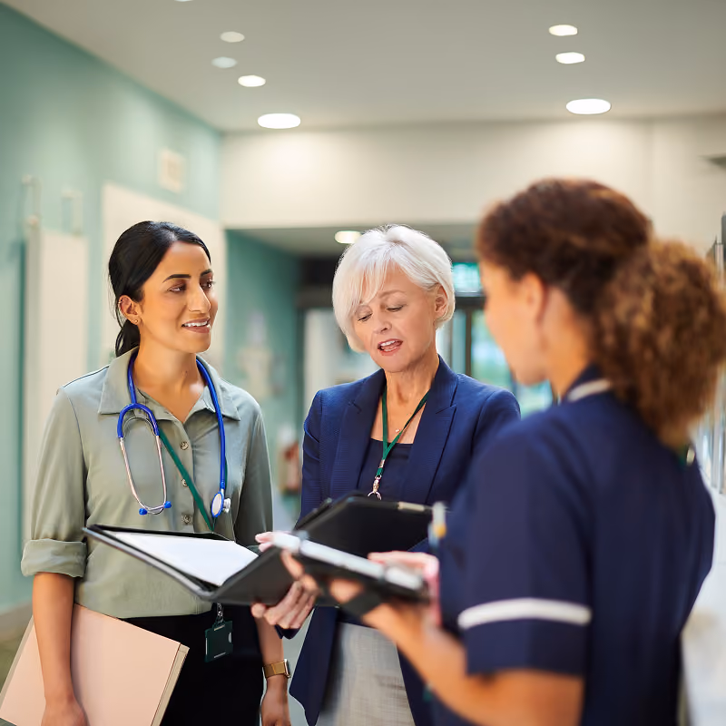 Three healthcare professionals standing in a hospital corridor, discussing information from clipboards and a tablet; one wears a stethoscope.