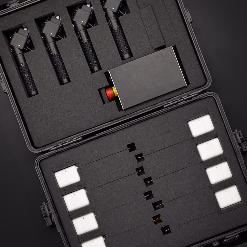 Open black hard case containing four black handheld devices with angled heads, a rectangular black electronic component with colorful parts, and eight rectangular white blocks secured with thin rods in foam compartments.