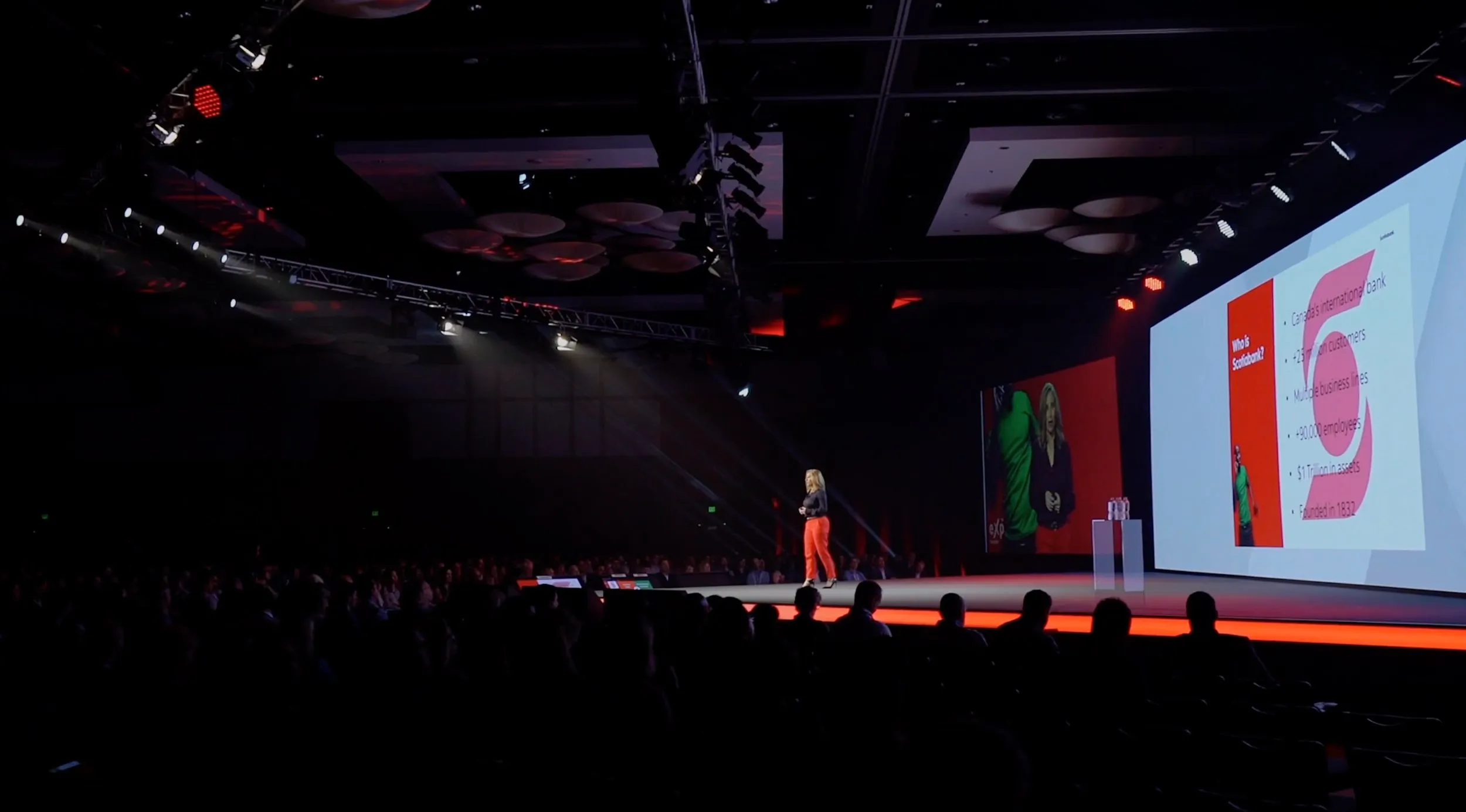 A woman speaker on a large stage in front of an audience presents a slide about Scotiabank with details on its international presence and business data, illuminated by stage lights in a dark conference hall.