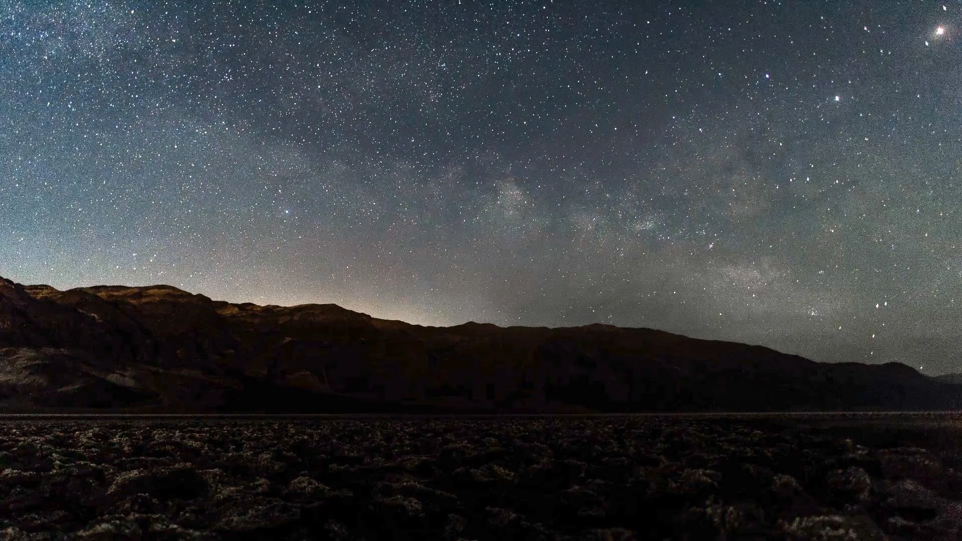Star-filled night sky with the Milky Way visible above dark mountain silhouettes and rocky terrain in the foreground.