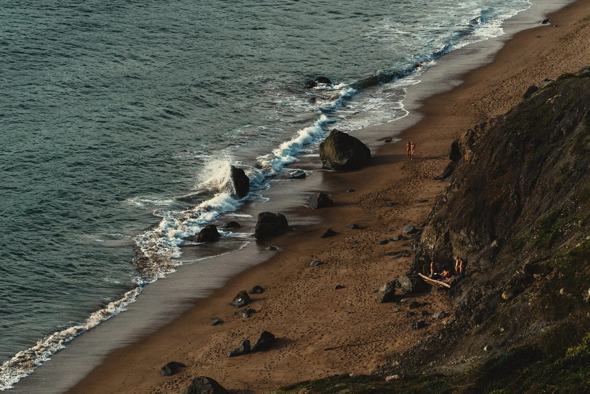 Sandy beach with large rocks, waves and people walking near a rocky cliff.