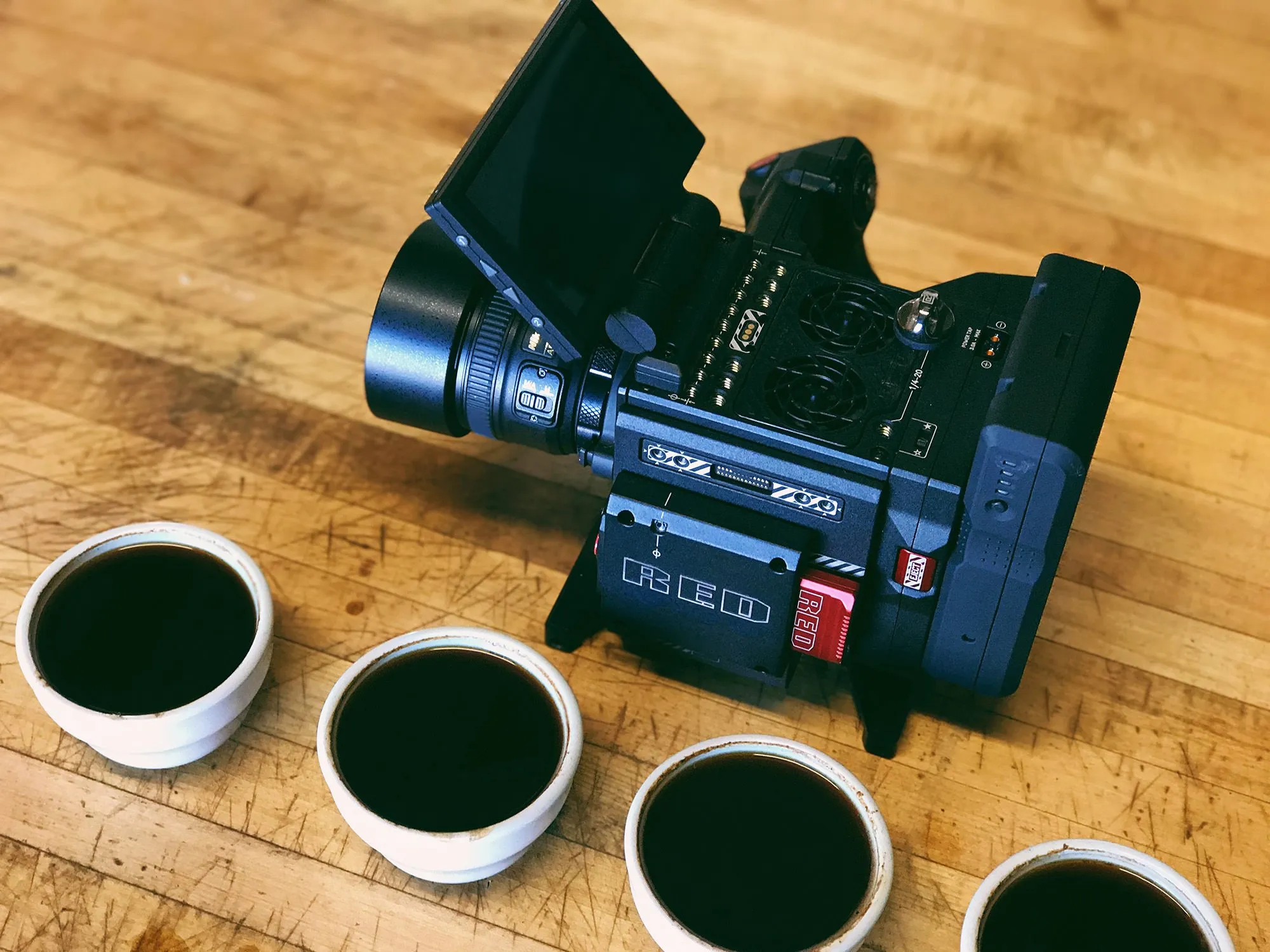 RED cinema camera on a wooden table beside four cups of black coffee.