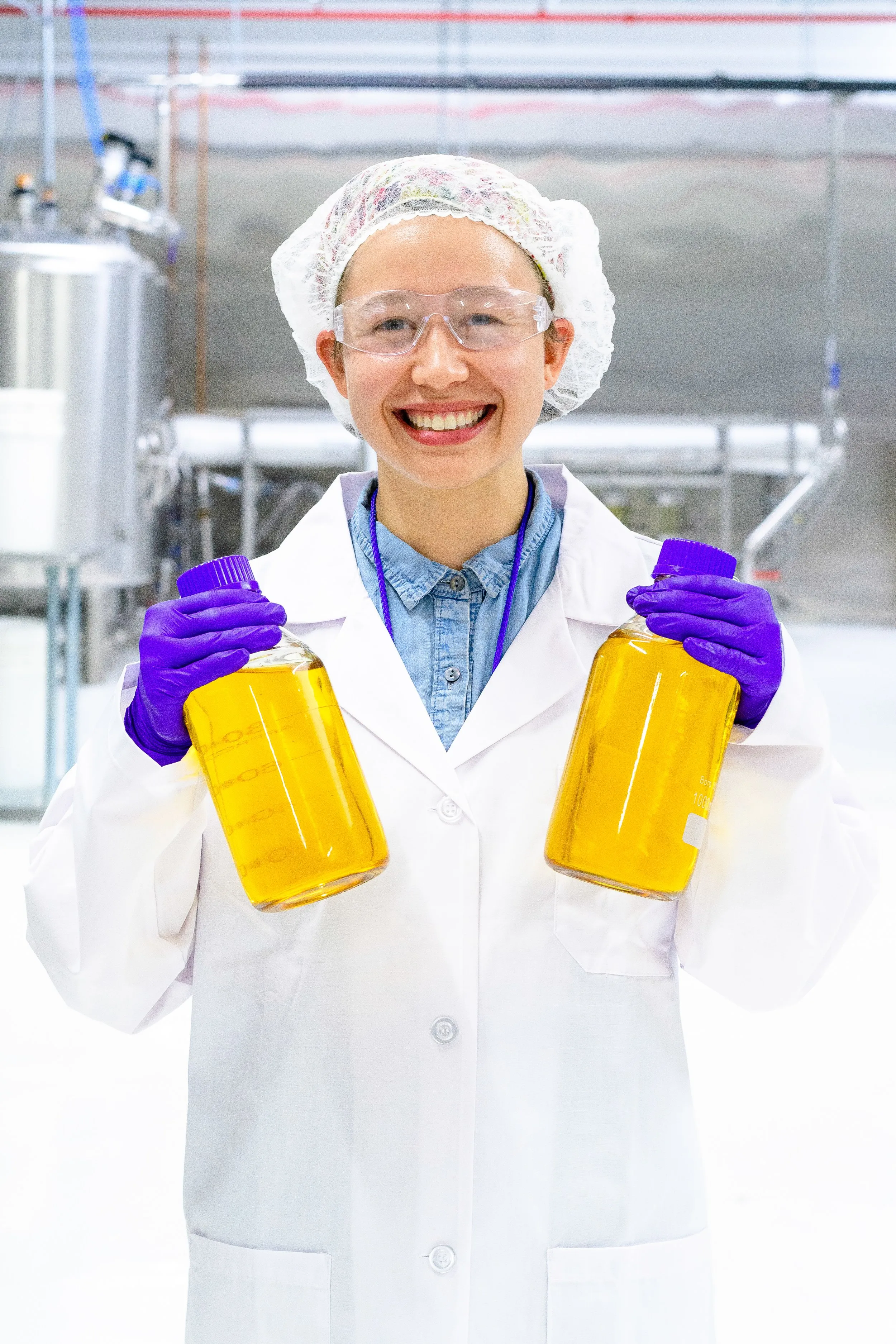 Smiling lab technician in safety glasses holding two large bottles of yellow liquid.