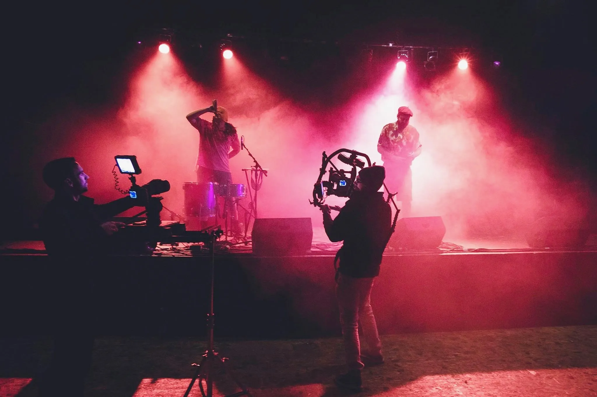 Two camera operators filming a singer and saxophonist on a smoky, pink-lit stage.