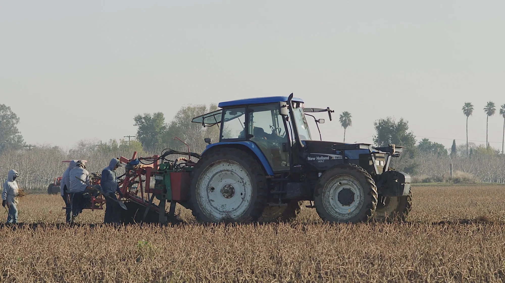Workers beside a blue New Holland tractor and farm equipment in a field.
