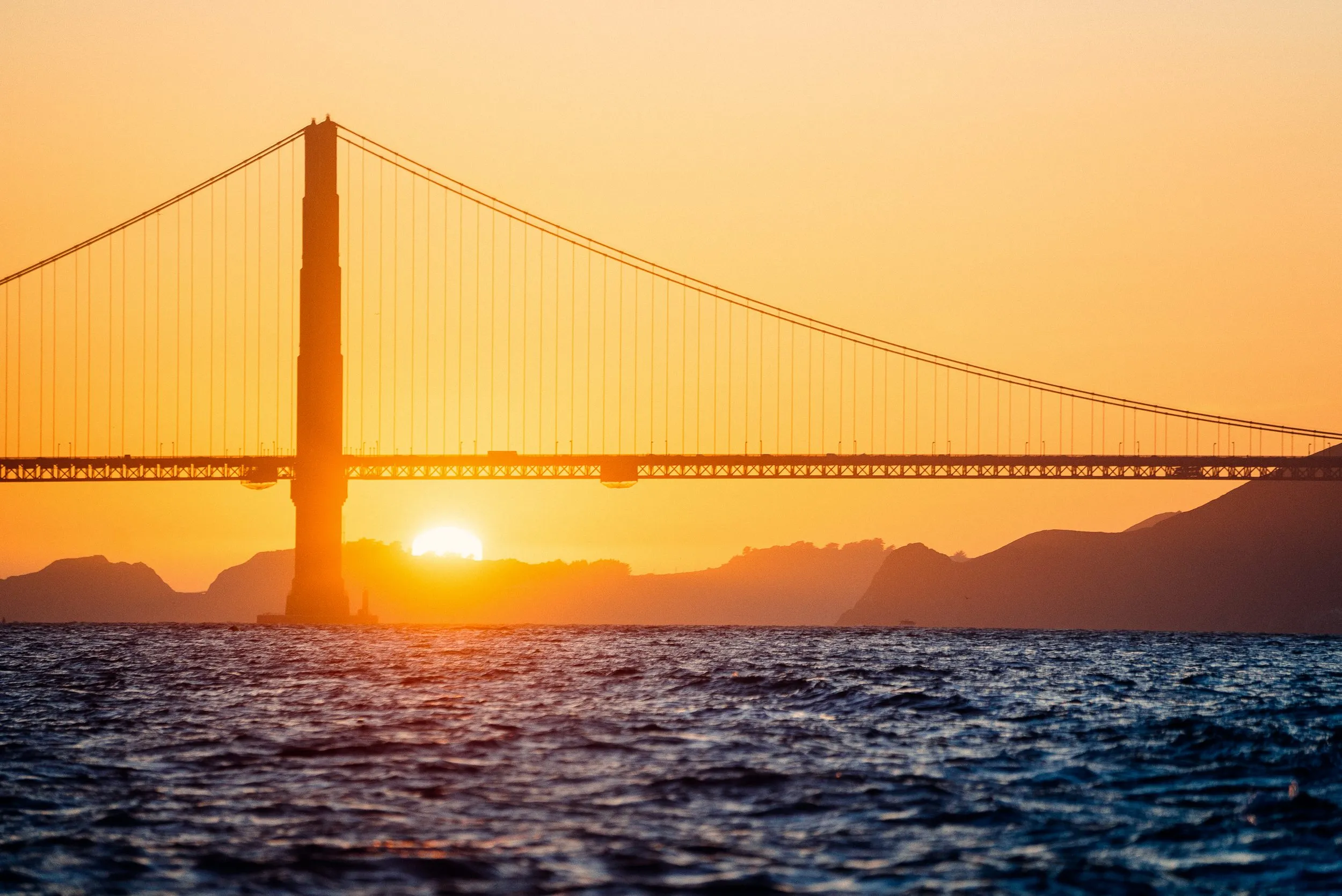 Golden Gate Bridge silhouetted at sunset over dark blue water.